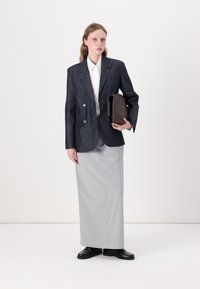 Blazer din denim închis cu butoane argintii, cămașă albă și fustă lungă gri. Modelul ține o poșetă maro și poartă pantofi negri.