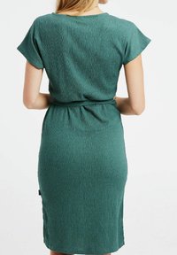 Teal mekko, joka on valmistettu teksturoidusta kankaasta, lyhyet hihat, tiukka muotoilu, vyötäröltä kapeutettu, polvipituinen, jossa on pieni sivutarra.
