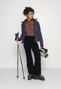 Tenue de ski comprenant une veste bleu marine, un haut marron et noir à motifs, un pantalon noir, des chaussures de ski, des bâtons de ski, et un casque avec un accent orange.