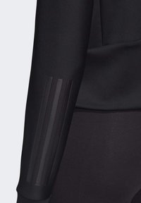 Gros plan sur un haut noir à manches longues avec un détail de rayures verticales en relief sur l'avant-bras, porté avec un pantalon noir ajusté.