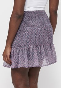 Femme portant une jupe courte taille haute violette à motif floral, avec une ceinture smockée et un ourlet volanté, associée à un haut blanc.