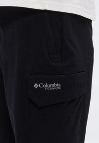 Μαύρο παντελόνι με πλαϊνή τσέπη cargo που φέρει το λογότυπο "Columbia Titanium", φορεμένο με λευκό πουκάμισο.