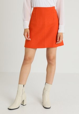 Jupe A-line orange en tissu texturé, dotée de poches avant et d'une taille haute. Associée à des bottes blanches à talons carrés montantes jusqu'au genou.