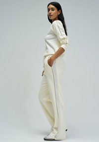 Femme se tenant de côté, portant un ensemble de loisirs couleur crème avec des accents argentés et des sneakers blanches, devant un fond gris clair uni.