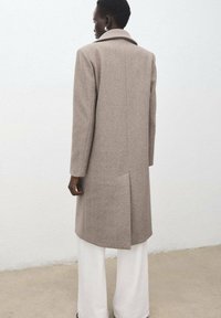 Cappotto di lana marrone chiaro con spacco posteriore, colletto a lancia e maniche lunghe. Indossato sopra pantaloni larghi bianchi su sfondo neutro.