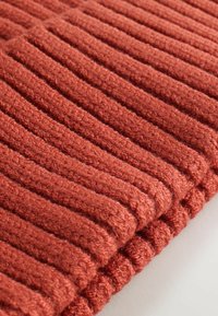 Tissu tricoté de couleur rouille avec une texture côtelée, présentant des lignes épaisses et surélevées. Le matériau semble doux et chaud, avec un aspect confortable et accueillant.