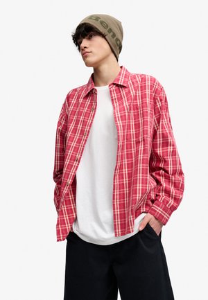 Ung person iført en grøn hue, rød ternet skjorte over en hvid T-shirt og sorte bukser, med hænderne i lommerne, ser til siden.