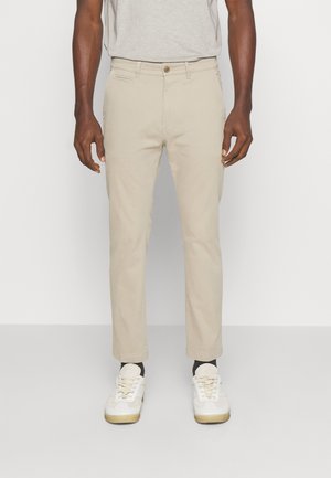 Mann trägt beige Slim-Fit-Hose, weiße Sneaker und dunkle Socken, steht vor einem einfarbigen hellen Hintergrund.