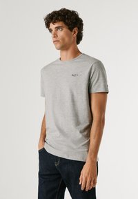 Mand med krøllet hår iført en lysegrå kortærmet t-shirt og mørke jeans, stående med den ene hånd i lommen og kigger let mod venstre.