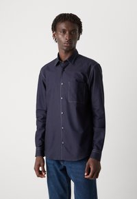 Navy button-up shirt with a classic collar, chest pocket, contrasting white stitching, and long sleeves, paired with blue denim jeans.