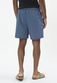 Costumi da bagno a strisce blu e bianche con cintura elastica e tasche laterali. Indossati con scarpe beige slip-on.