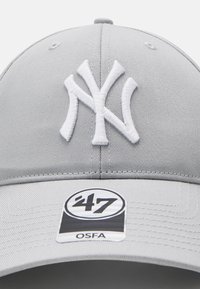 Casquette de baseball grise avec un logo NY brodé blanc bien en vue, une visière courbée et une étiquette de taille indiquant ‘47 OSFA. Tissu texturé.