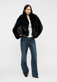 Manteau en fausse fourrure noire à larges manches, porté sur une chemise blanche, associé à un jean taille haute bleu et des escarpins noirs à bout pointu.