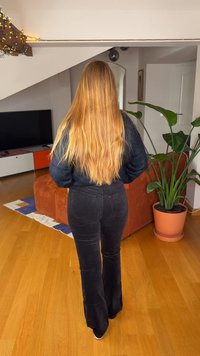 Schwarze ausgestellte Hose und ein schwarzer Pullover werden getragen, mit langem, welligem Haar, das den Rücken hinunterfällt. Im Hintergrund steht eine Topfpflanze.