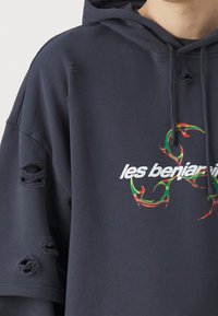 Felpa nera over size con dettagli scuciti e un logo grafico colorato al centro. Il materiale sembra morbido e texturizzato.