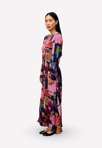 Frau mit langen schwarzen Haaren steht in einem bunten, bodenlangen Patchworkkleid mit Mustern von Pflanzen und Möbeln, trägt schwarze Ballerinas.