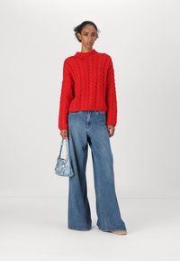 Rød kabelstrikket genser, løse høywaisted denimjeans, lyseblå håndveske med kjededetalj. Modellen står mot en enkel hvit bakgrunn.
