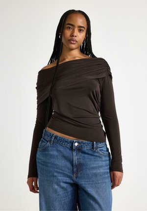 Kvinde med lange fletninger iført en sort off-shoulder bluse med lange ærmer og blå højtaljede jeans mod en ensfarvet lys baggrund.