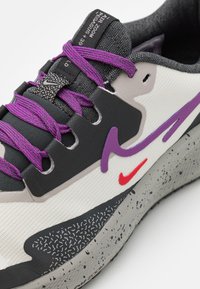 Sportstrumpor med en vit mesh-övre, svarta accenter, lila snören och en texturerad grå sula med svarta fläckar. Röd logodetalj närvarande.