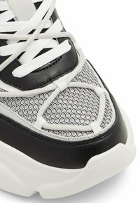 Sneaker in bianco e nero realizzato in materiali in rete e pelle, con un motivo testurizzato, lacci piatti e una suola bianca spessa.