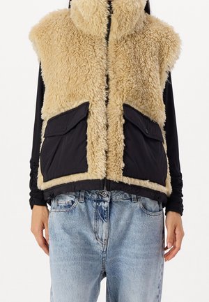 Veste beige sans manches en fausse peau retournée avec poches à rabat noires, portée sur un haut noir à manches longues et un jean bleu clair.