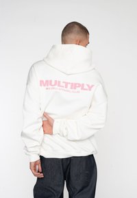 Sudadera blanca con gran texto rosa "MULTIPLICAR" en la parte posterior, con un corte relajado y capucha con cordón. Tela texturizada, mangas con puños.