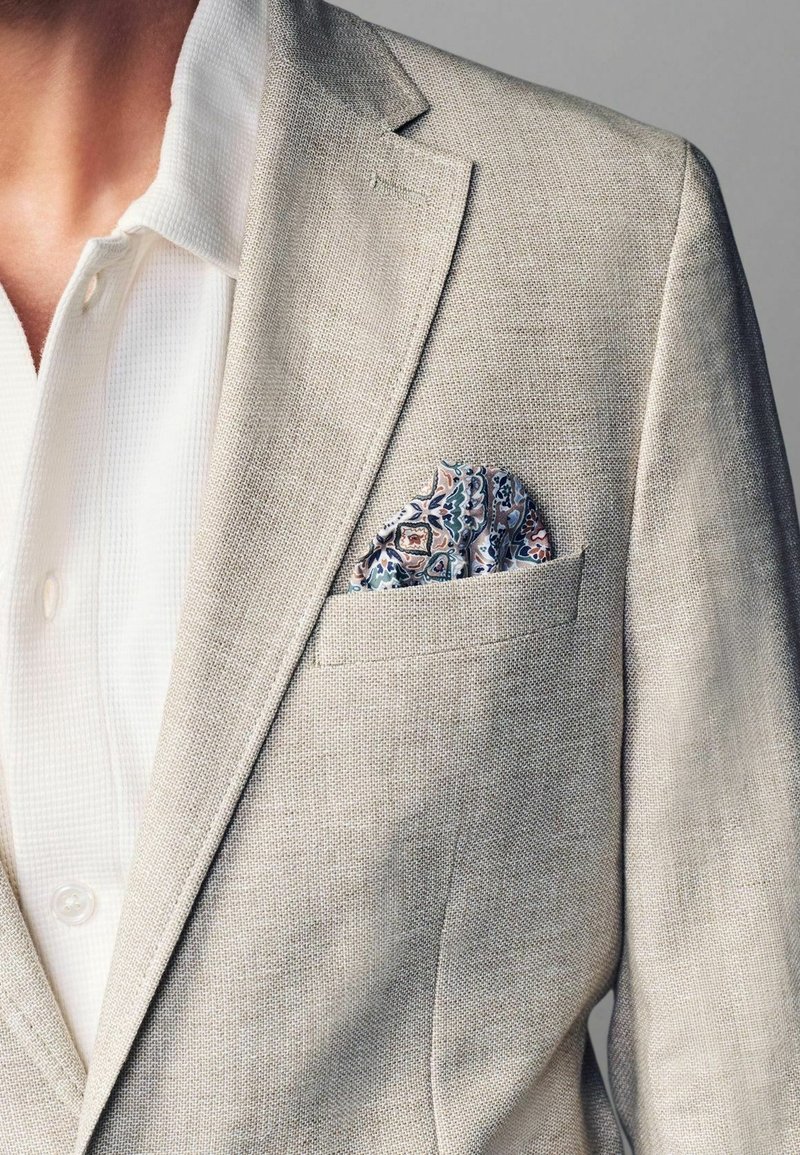 Lys beige linned blazer med klapkrave, parret med en hvid struktureret skjorte. Har en paisley lommefyr i blå, pink og grønne nuancer.