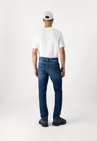Vaqueros de hombre de corte recto en azul, fabricados en denim, con diseño estándar de cinco bolsillos y una etiqueta morada en la parte trasera de la cintura. Combinados con una camiseta blanca.