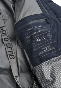 Mörkblå och grå ytterkläder med en texturerad nedre panel, broderade logotyper, vård symboler och ett tygmärke med produktinformation.