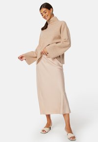 Beige pletený svetr s vysokým krkem a širokými rukávy, kombinovaný s světle béžovou saténovou midi sukni a bílými sandály na podpatku.