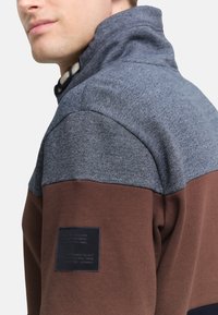 Fleecepullover mit hohem Kragen, in Grau- und Brauntönen, mit strukturierter Stoffoberfläche und einem schwarzen Markenetikett am Ärmel.