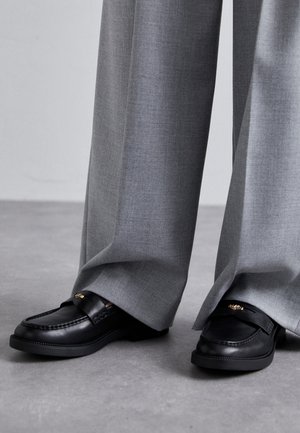 Mocassins en cuir noir à bout rond, avec des accents en métal doré, associés à un pantalon large en tissu texturé de couleur grise.