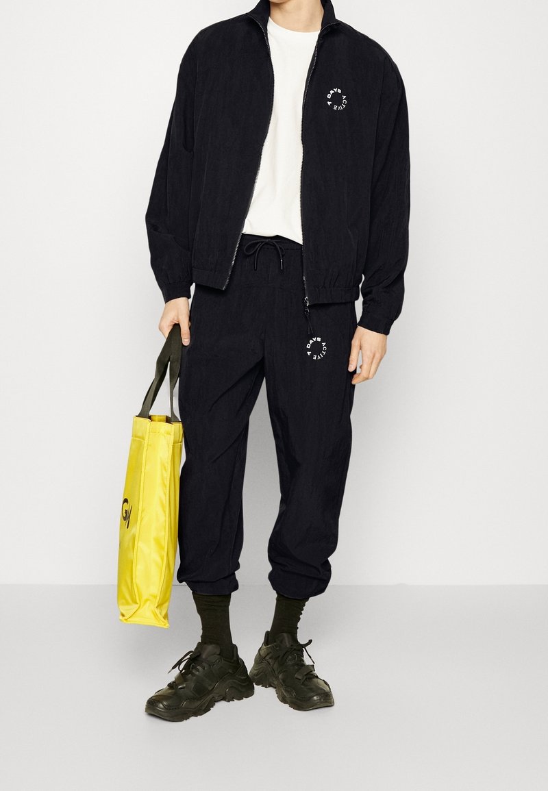Chaqueta negra con cremallera y pantalones a juego, hechos de tejido ligero. Incluye detalle del logo en blanco. Sostiene una bolsa de tela amarilla con texto negro.