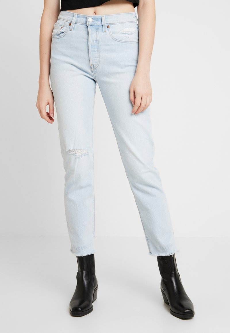 Femme portant un jean taille haute bleu clair déchiré, ourlets effilochés, bottines noires et un top noir court sur un fond neutre.