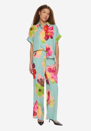 Kobieta stojąca w jasnoniebieskiej, kwiatowej koszuli na krótki rękaw z guzikiem oraz pasujących szerokich spodniach, uzupełnionych czarnymi szpiczastymi szpilkami.