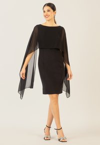 Schwarzes Kleid mit einem fließenden, transparenten Überzug, figurbetontem Schnitt, rundem Halsausschnitt und verzierten Sandalen mit Schleifen. Glatte Textur, elegantes Design.