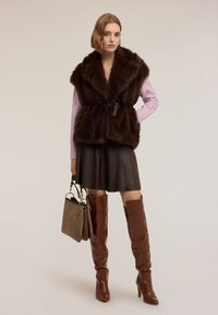 Brązowa futrzana kamizelka z szerokim kołnierzem, taliowana paskiem, w zestawieniu z lawendowym swetrem, czarną skórzaną spódnicą i brązowymi kozakiem za kolano.