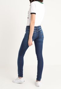 Vaqueros ajustados en azul oscuro, con una cintura de altura media, diseño de cinco bolsillos y acentos de costura mínimos. Combinados con zapatillas blancas.