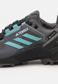 Zapatilla deportiva negra y gris con acentos en turquesa, parte superior de malla texturizada, marca Gore-Tex, patrón de banda de rodadura agresivo en la suela y refuerzos reforzados.