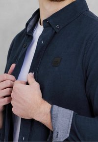 Námořnická manšestrová košile s límečkem na knoflíky, s šedými flanelovými manžetami a černou logo nášivkou nad kapsou. Hladká textura.