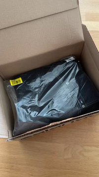 Διπλωμένα μαύρα ρούχα τυλιγμένα σε πλαστικό μέσα σε ανοιχτό χαρτονένιο κουτί αποστολής πάνω σε ξύλινο πάτωμα.