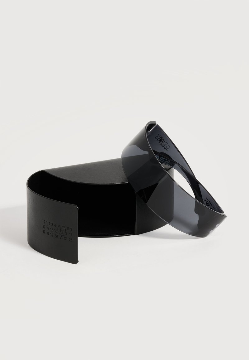 Ceinture large en cuir noir avec une grande boucle en plastique brillante et incurvée, posée sur une surface blanche avec des marques de designer visibles.