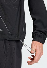 Μαύρο ελαφρύ αθλητικό μπουφάν με τσέπη με φερμουάρ, ελαστικά μανίκια και κορδόνι στη μέση για ρυθμιζόμενη εφαρμογή. Υφή σχεδιαστικών λεπτομερειών.