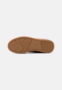 Semelle de chaussure avec grip en caoutchouc marron présentant des motifs de bande de roulement circulaires, en zigzag et rayés, ainsi que du texte de marque en relief sur un fond blanc.