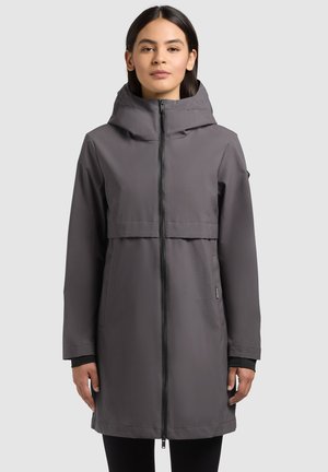 Femeie purtând o haină lungă, gri, cu glugă și fermoar, stând pe un fundal simplu, gri deschis.