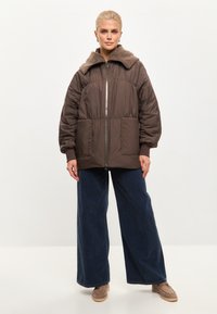 Bruine pufferjack met een pluche, tan-kleurige kraag, ritssluiting aan de voorkant, twee grote voorkezen en een oversized pasvorm, gecombineerd met wijde denimbroeken.