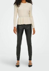 Cremefarbener, gerippter Peplum-Pullover mit langen Ärmeln, kombiniert mit schwarzen, enganliegenden Hosen aus Kunstleder und schwarzen spitzen Schuhen mit Kettenakzenten.