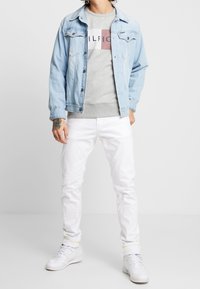Giacca di jeans azzurro chiaro sopra un sudario grigio con un logo colorato, abbinata a jeans bianchi e sneakers bianche, su uno sfondo neutro.