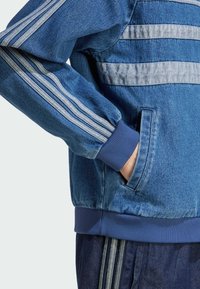 Blå denimjacka med grå accentstripes, ribbad mudd och sidofickor. Har en avslappnad passform och texturerad tyg.