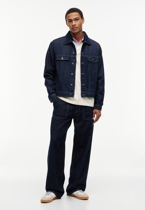 Ung mand iført mørkeblå denimjakke og bukser, hvid skjorte under, og hvide sneakers med brune såler, stående.